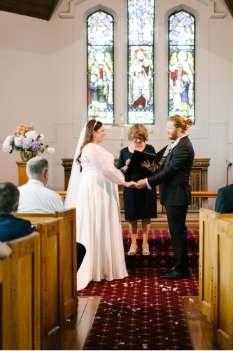 Bride, groom exchanging vows in a church with a stained glass window in the background.